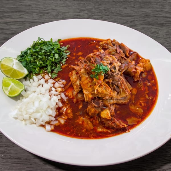 Birria de chivo, a goat stew, with cilantro, diced onions, and lime wedges on a white plate.