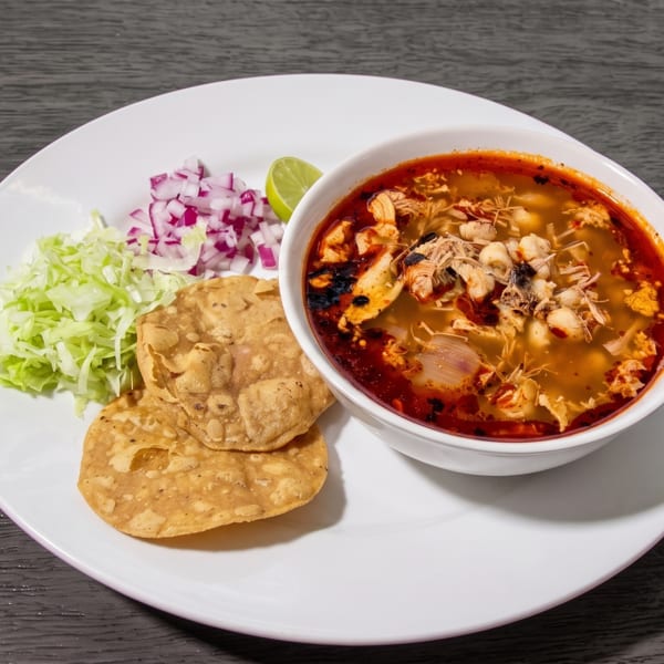 A bowl of pozole with shredded chicken, hominy, and red chili oil, served with tostadas and garnishes.