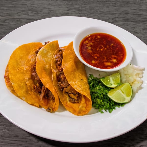 Four golden-fried tacos with meat filling, a side of red sauce, cilantro, diced onions, and lime wedges.