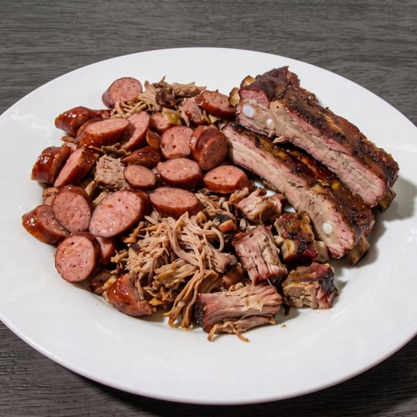 A white plate piled high with sliced sausage, pulled pork, and two racks of ribs.